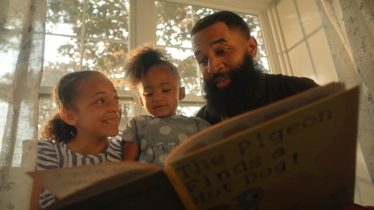 A dad reading a picture book to his two daughters by a sunlit window, from the Grown Up Dad trailer
